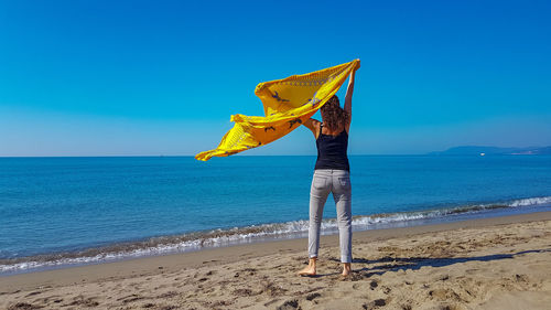 Rear view of woman holding textile at beach against clear sky