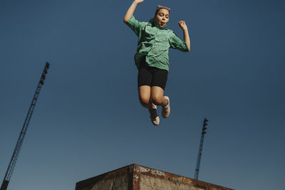 Carefree girl making face while jumping in mid-air against blue sky
