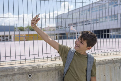 Smiling boy taking selfie through mobile phone while standing against wall