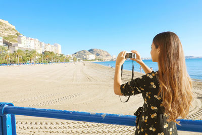 Woman photographing at beach against clear blue sky