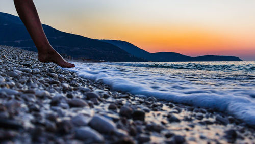 Low section of person standing on beach against sky during sunset