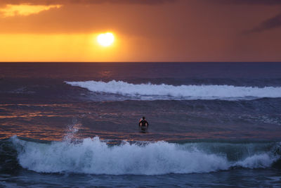 Surfer in sea against cloudy sky during sunset