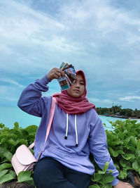 Portrait of woman wearing hijab holding photographs while sitting against sea