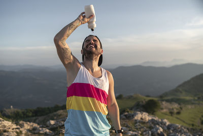 Athlete pouring water on face while standing on mountain against clear sky