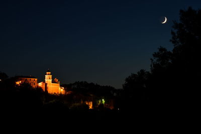 Silhouette trees and illuminated buildings against sky at night