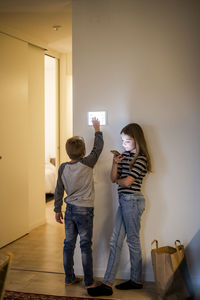 Brother using digital tablet mounted on wall while sister text messaging on mobile phone in smart home