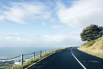 Empty road by sea against cloudy sky