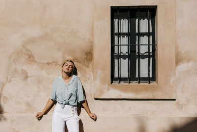 Beautiful woman with eyes closed standing against beige wall during sunny day