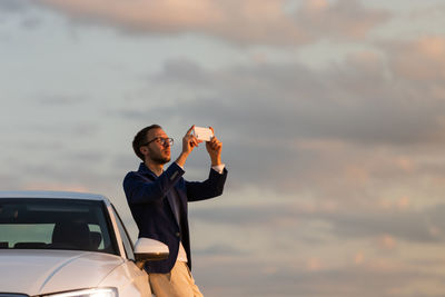 Man standing on car against sky during sunset