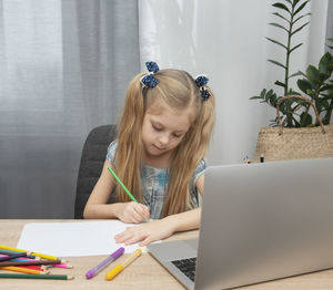 Girl drawing over paper by laptop on table