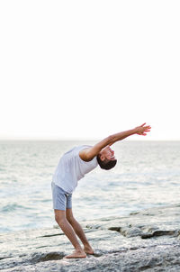 Side view of man exercising while standing on beach against clear sky