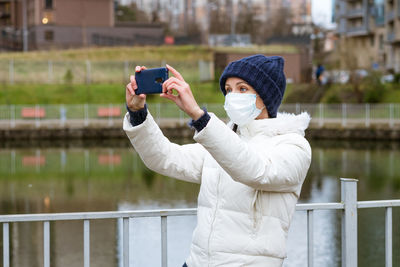 Young woman in a protective mask in a jacket takes pictures on the phone