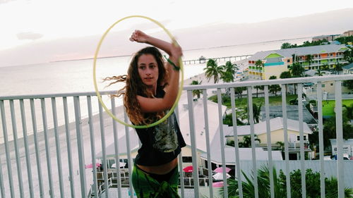 Young woman playing with plastic hoop by railing at beach against sky