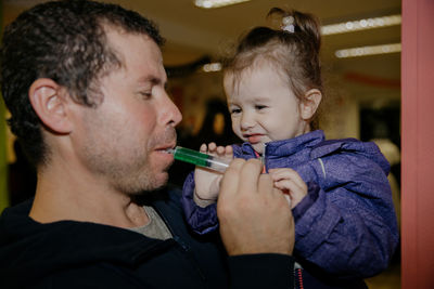 A father with his daughter in his arms drinks liquid from a syringe.