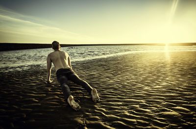 Shirtless man exercising at beach against sky during sunset