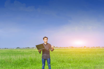 Portrait of man holding laptop while standing on field against sky