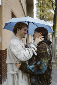 Side view of young romantic gay couple standing under umbrella at street