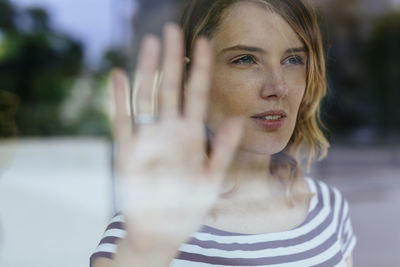 Portrait of young woman looking through window touching glass with her hand