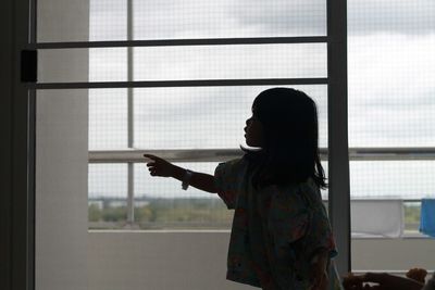 Side view of girl pointing while standing by glass window