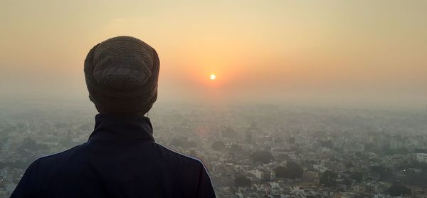 Rear view of man looking at cityscape against sky during sunset