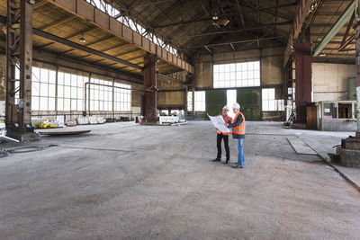 Two men with plan wearing safety vests talking in old industrial hall