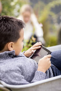 Boy using digital tablet while sitting in wheel barrow with mother gardening in background