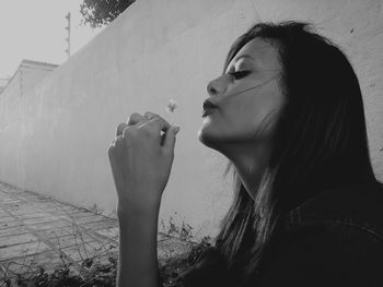 Close-up of young woman blowing flower by wall