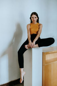 Full length portrait of young woman sitting by wall