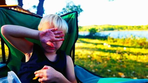 Boy with hand covering eyes relaxing on seat at field