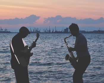 Musician playing musical instruments while standing against sea during sunset