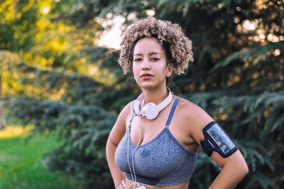 Young ethnic curly haired sportswoman with smartphone on shoulder holder and headphones on neck standing against green trees and looking at camera during training in park