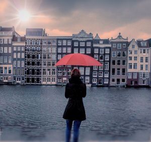 Rear view of mid adult woman with umbrella while standing by river against cloudy sky during sunset