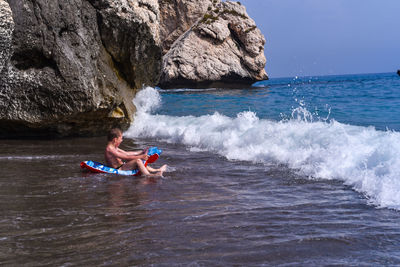 Boy sitting on inflatable raft at beach against sky