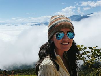 Smiling woman wearing sunglasses and knit hat standing on mountain during foggy weather
