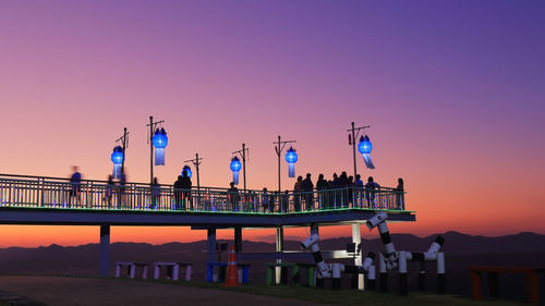 People at illuminated bridge against clear sky during sunset