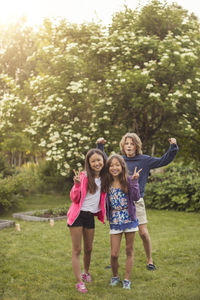 Portrait of happy sisters showing peace sign while friend flexing muscle on grass in back yard