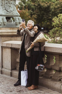 Happy senior couple taking selfie through smart phone while standing on bridge