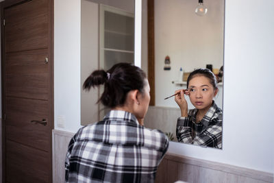 Focused charming ethnic female applying eyebrow pencil while doing makeup and looking in mirror