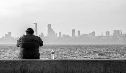 Rear view of man sitting on retaining wall by sea at marine drive