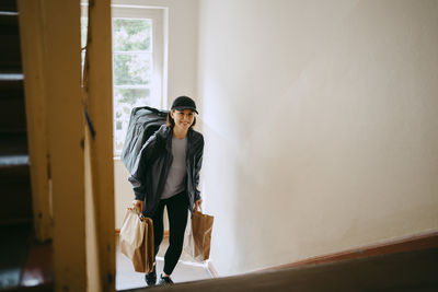 Smiling female delivery person holding bags and moving up on building staircase