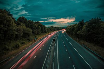 Light trails on road amidst trees against cloudy sky at dusk