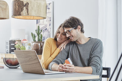 Smiling couple with a card using laptop on table at home