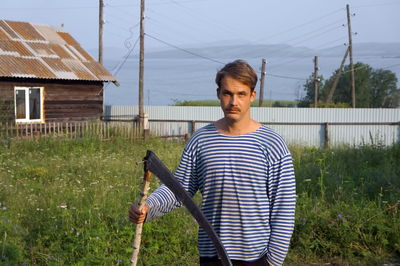 A mustachioed men in a striped vest stands with a scythe in his hands in the courtyard of a village