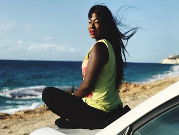 Side view of woman sitting on car at beach against sky