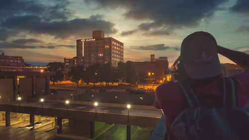 Woman looking at illuminated cityscape against sky at sunset