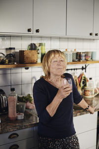 Blond senior woman with eyes closed holding drinking glass in kitchen at home