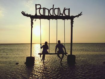 Silhouette couple sitting on swing in sea during sunset