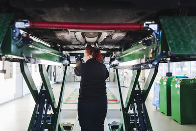 Rear view of female mechanic standing under car on hydraulic lift