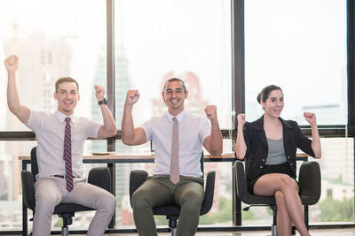 Happy business people celebrating success while siting on chairs against window at office