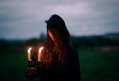 Young woman holding candles while standing outdoors against sky at sunset
